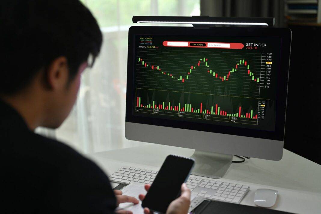 Young Male Financial Analyst Using Mobile Phone And Monitoring Stocks Data Candle Charts On Screen