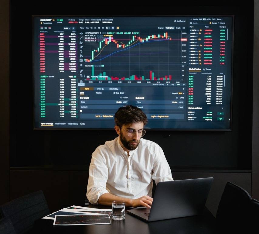 a person studying their laptop with a large monitor in the back showing a crypto stock data.
